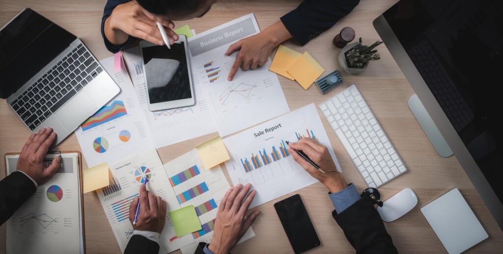 A top-down view of a group of professionals collaborating at a desk covered with business reports, charts, a laptop, a tablet, sticky notes, and a keyboard. They are pointing and discussing data visualizations on printed sheets.
