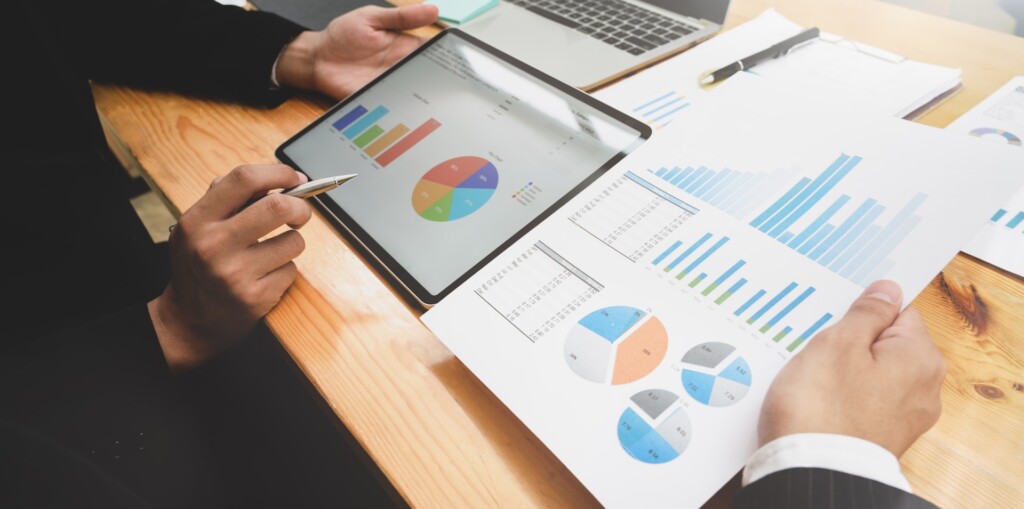 Two people reviewing financial and business reports with colorful graphs and pie charts on printed documents and a tablet at a wooden desk. One person is holding the paper while the other is pointing at the tablet screen with a pen.