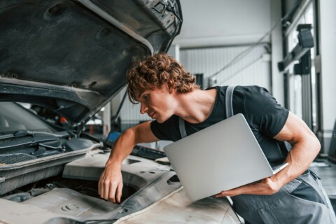 Technician using a laptop to diagnose and repair a car engine in a modern workshop, highlighting innovation and startup-driven repair services.