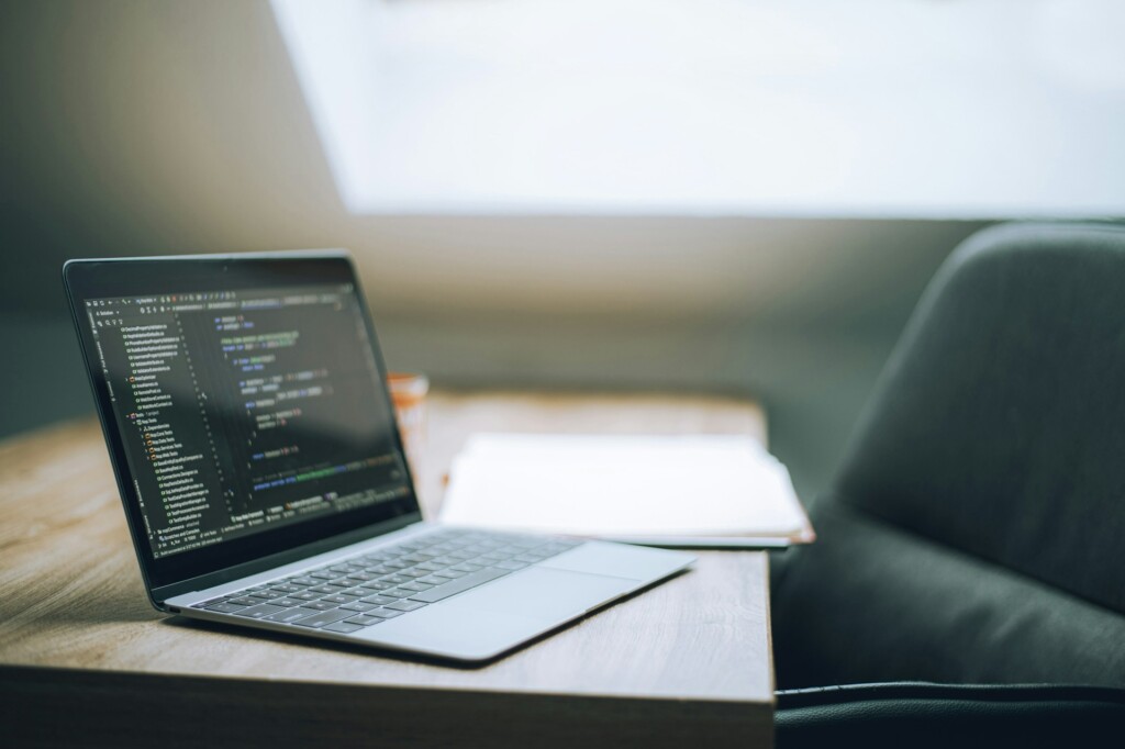 Laptop screen showing code on a wooden desk with notebook and coffee cup in a bright modern workspace, representing startup founder productivity with SaaS tools.​