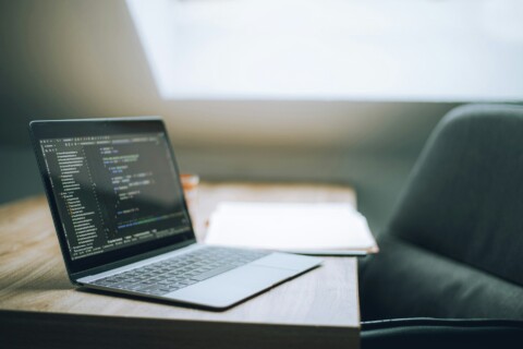 Laptop screen showing code on a wooden desk with notebook and coffee cup in a bright modern workspace, representing startup founder productivity with SaaS tools.​