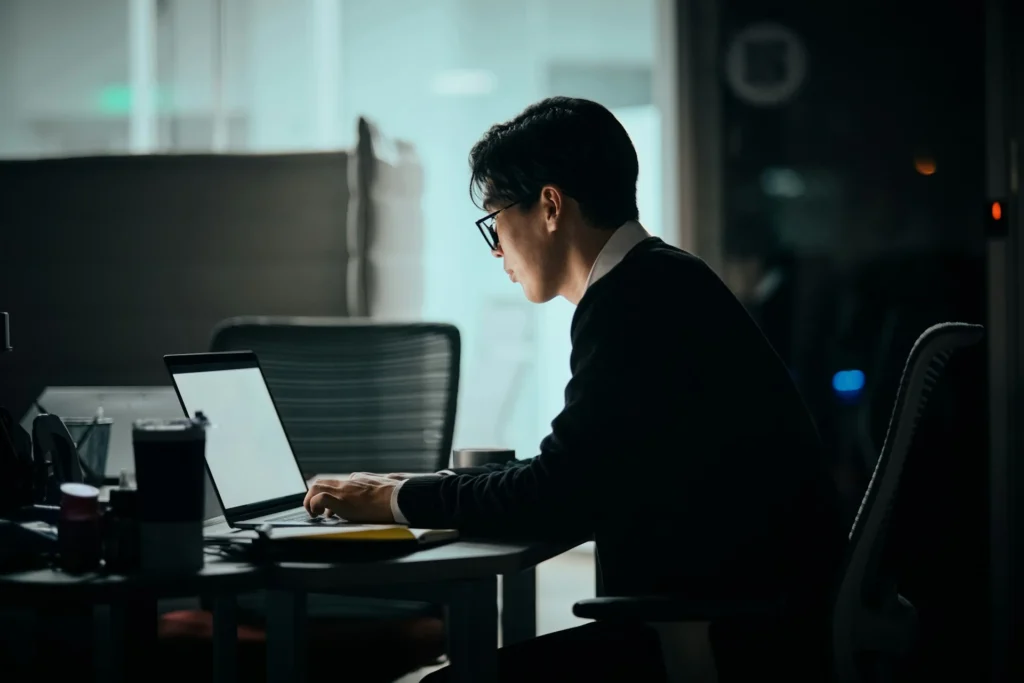 A man wearing glasses and a black sweater sits at a desk working on a laptop in a dimly lit office at night, viewed from a side profile angle."