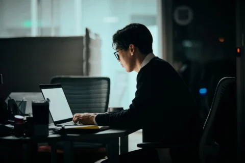 A man wearing glasses and a black sweater sits at a desk working on a laptop in a dimly lit office at night, viewed from a side profile angle."